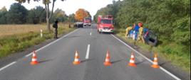 w rowie po prawej stroni drogi stoi niebieski samochód a przy nim trzech strażaków parę metrów dalej po lewej stronie drogi leży auto na dachu na drodze stoją dwa wozy strażackie