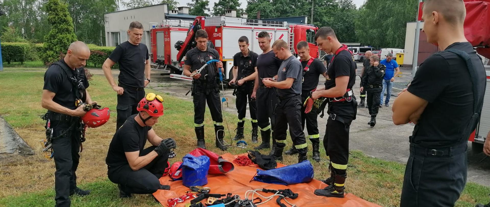 Strażacy stoją nad matą, na którym rozłożony jest sprzęt wysokościowy i omawiają przebieg ćwiczeń. W tle widać samochody strażackie.
