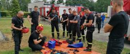 Strażacy stoją nad matą, na którym rozłożony jest sprzęt wysokościowy i omawiają przebieg ćwiczeń. W tle widać samochody strażackie.