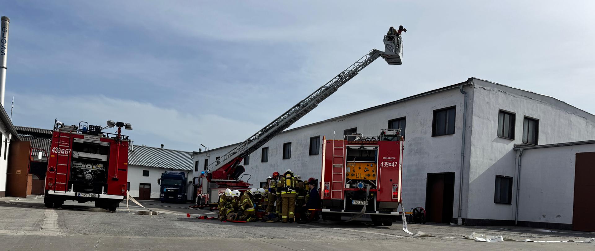 Zastępy stoją na placu zakładu. Strażacy prowadzą działania.