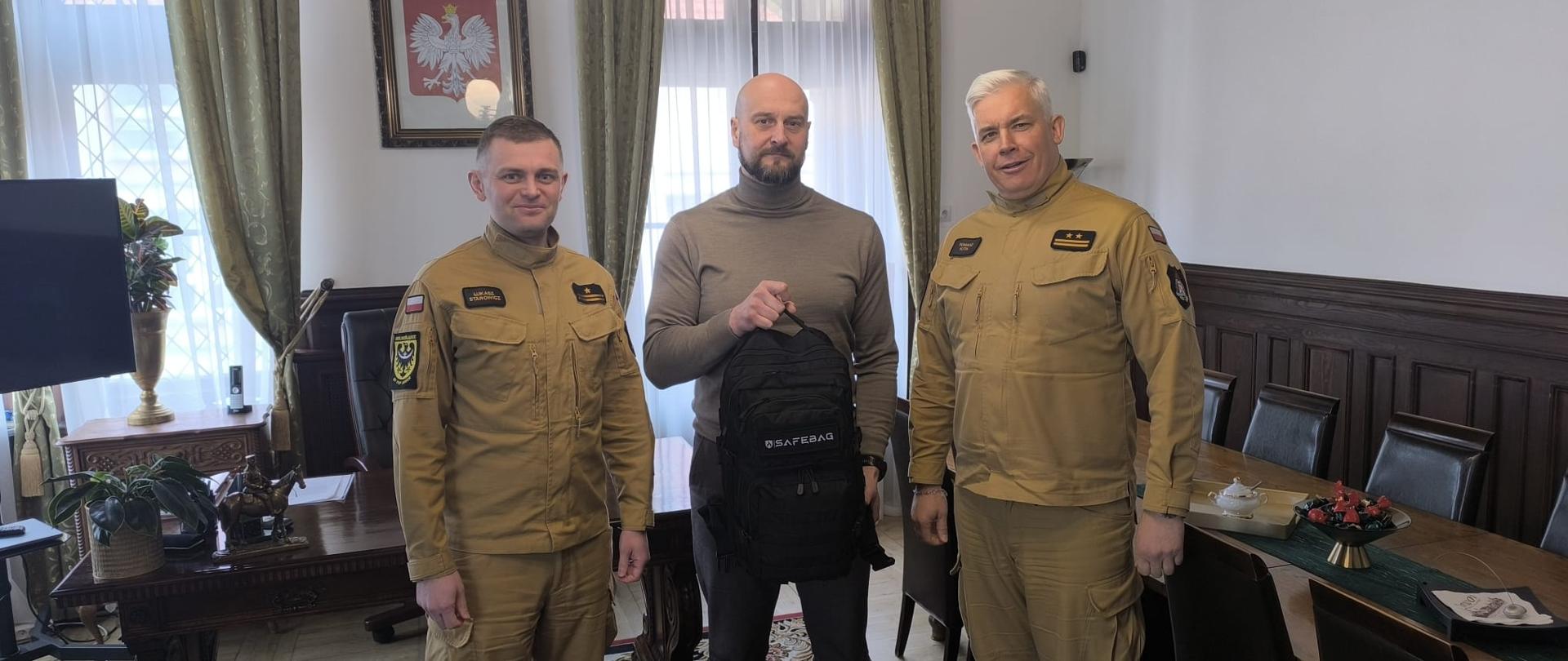 Zastępca Burmistrza przekazuje plecak ewakuacyjny na ręce Komendanta Powiatowego PSP w Jaworze. Osoby w gabinecie burmistrza