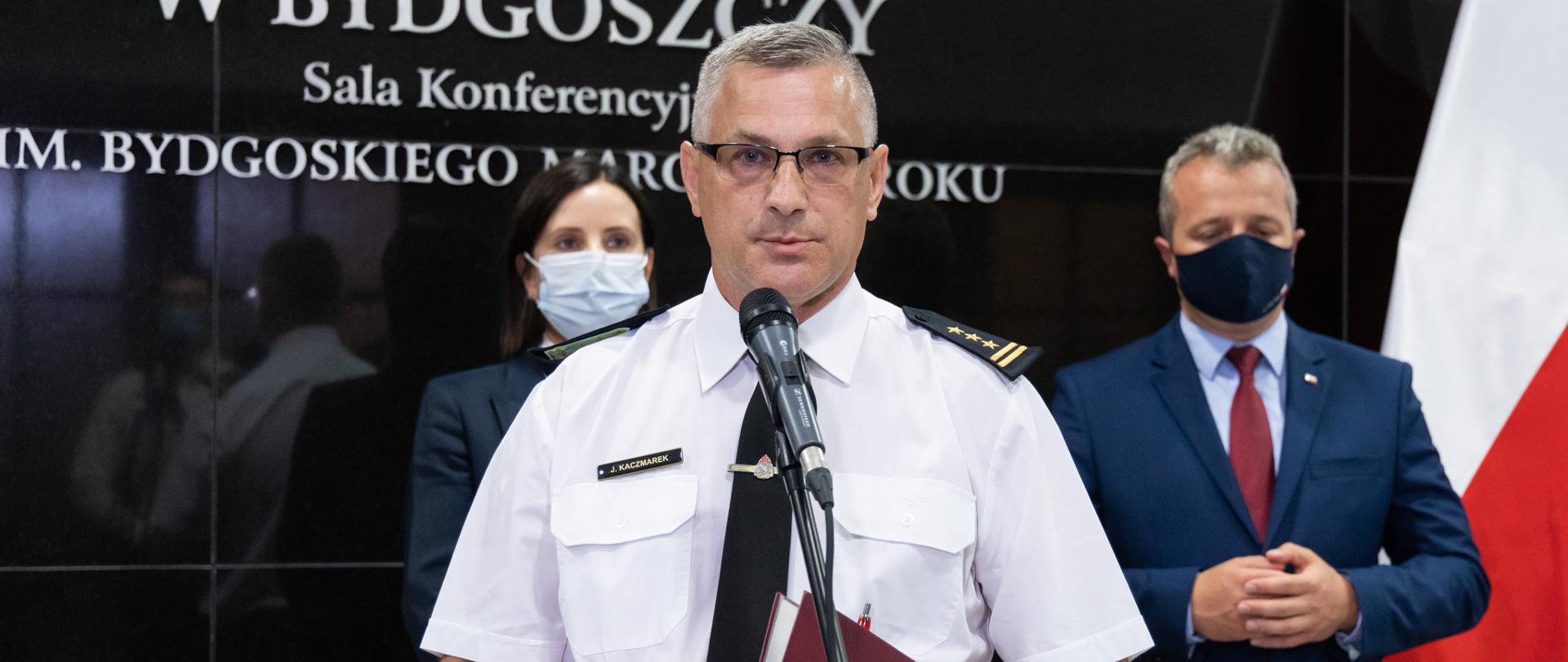 Na pierwszym planie komendant wojewódzki PSP przy mikrofonie, w umundurowaniu wyjściowym - letnim. Za nim kurator oświaty i wojewoda. W tle tablica Urzędu Wojewódzkiego i flagi narodowe. Trwa konferencja z mediami.
