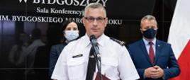 Na pierwszym planie komendant wojewódzki PSP przy mikrofonie, w umundurowaniu wyjściowym - letnim. Za nim kurator oświaty i wojewoda. W tle tablica Urzędu Wojewódzkiego i flagi narodowe. Trwa konferencja z mediami.