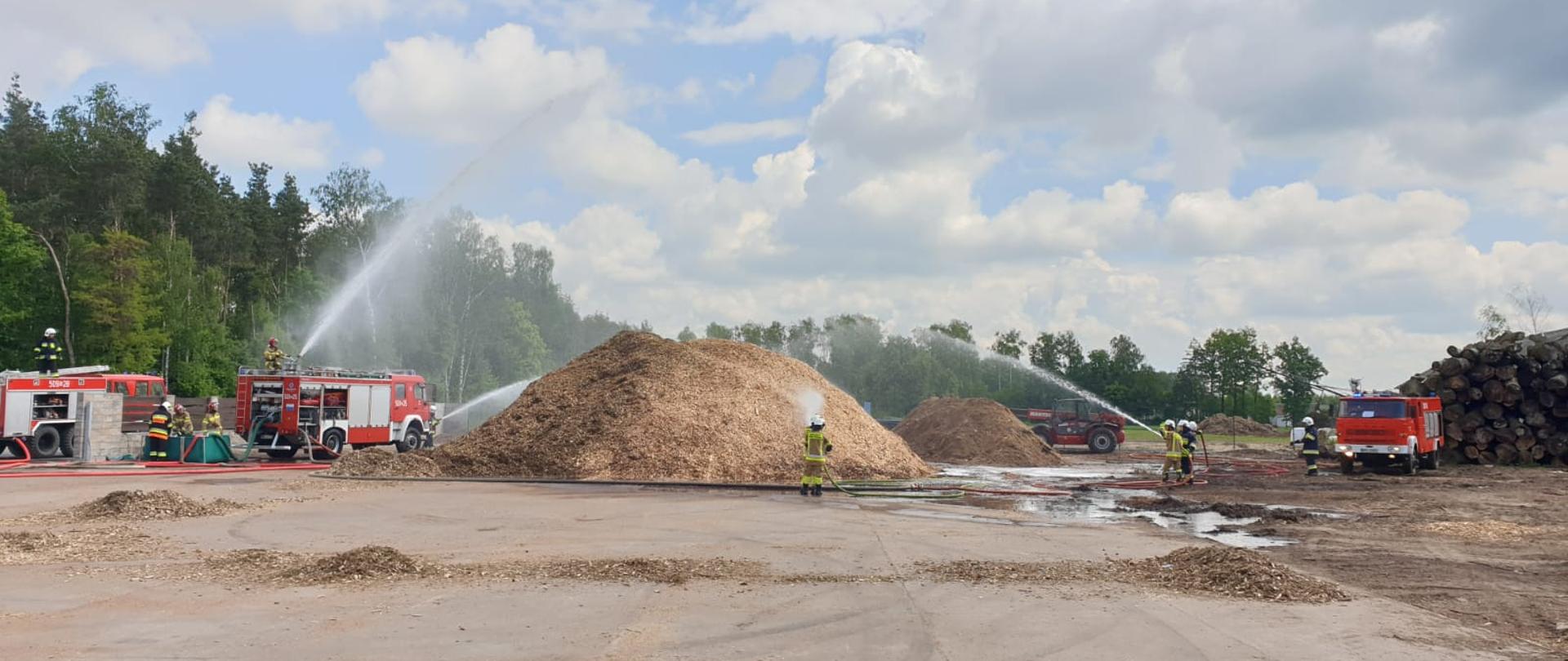 Strażacy podają cztery prądy wody na pryzmę zrębki. Woda podawana jest z samochodów gaśniczych.