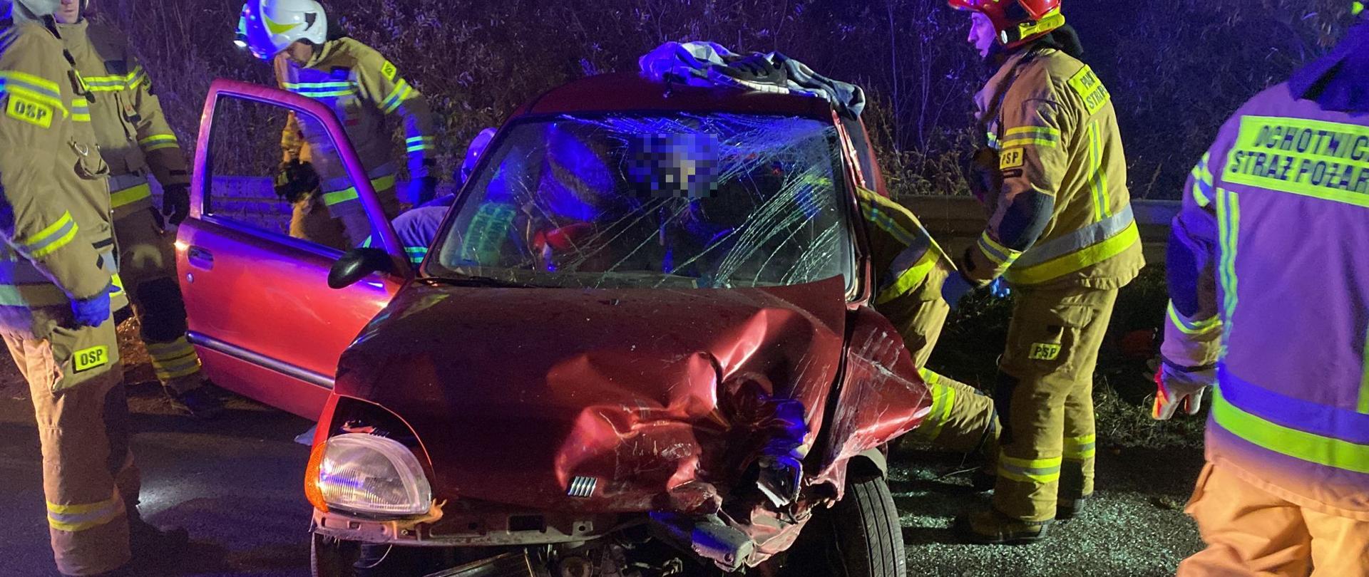 czerwony samochód z rozbitym przodem oraz bokiem od strony kierowcy wokół samochodu pięciu strażaków jeden strażak wchodzi do środka samochodu