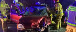 czerwony samochód z rozbitym przodem oraz bokiem od strony kierowcy wokół samochodu pięciu strażaków jeden strażak wchodzi do środka samochodu
