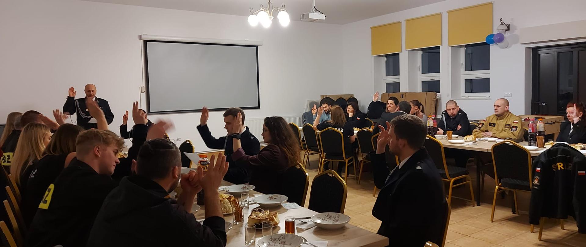 Na zdjęciu widać komendanta powiatowego PSP w Ustrzykach Dolnych mł. bryg. Emila Szmyda w towarzystwie druhów OSP Łodyna na spotkaniu sprawozdawczym. Wszyscy siedzą przy stole na którym stoją napoje ciastka paluszki oraz szklanki talerze i sztućce. 