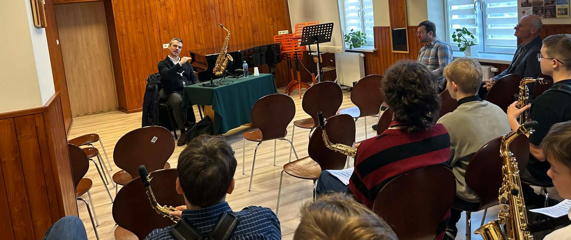 Prowadzący warsztaty siedzi przy stoliku z saksofonem, omawiając zagadnienia gry na instrumencie; przed nim siedzą uczniowie z saksofonami w sali szkolnej z drewnianymi ścianami i dużymi oknami.