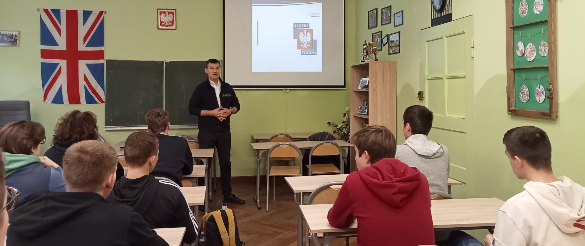 sala lekcyjna, wypełniona uczniami. Inspektor stojący przy tablicy szkolnej omawia slajd wyświetlany z rzutnika. Na slajdzie widać logo Służby Cywilnej.