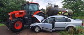 Na zdjęciu widać zderzenie dwóch pojazdów, samochodu osobowego i ciągnika rolniczego oraz strażaków