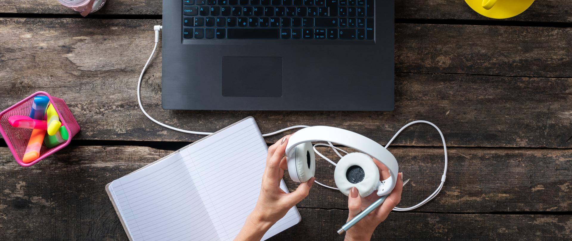 Zdjęcie (ujęcie od góry): widok drewnianego blatu, na który widać laptopa. Przed komputerem widoczne damskie dłonie trzymające białe słuchawki, obok notes i kolorowe zakreślacze.