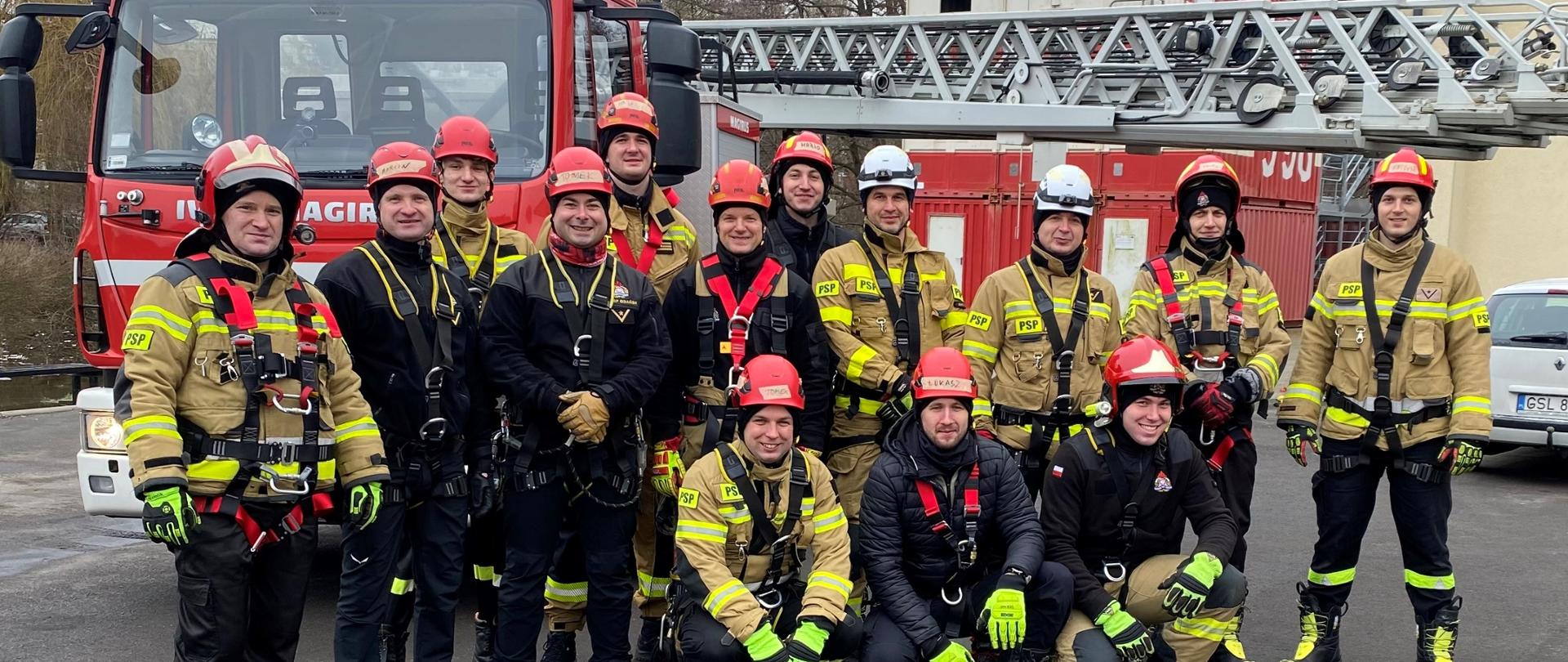 Strażacy w umundurowaniu specjalnym w hełmach oraz sprzęcie wysokościowym stoją obok siebie za nimi ustawiony jest samochód specjalny drabina.