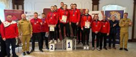 Kolorowa fotografia wykonana w pomieszczeniu. Przedstawia strażaków w strojach sportowych (dresach) trzymających dyplomy i medale. Część z nich jest na podium, a część poza nim. Komendanci w umundurowaniu służbowym są obok.