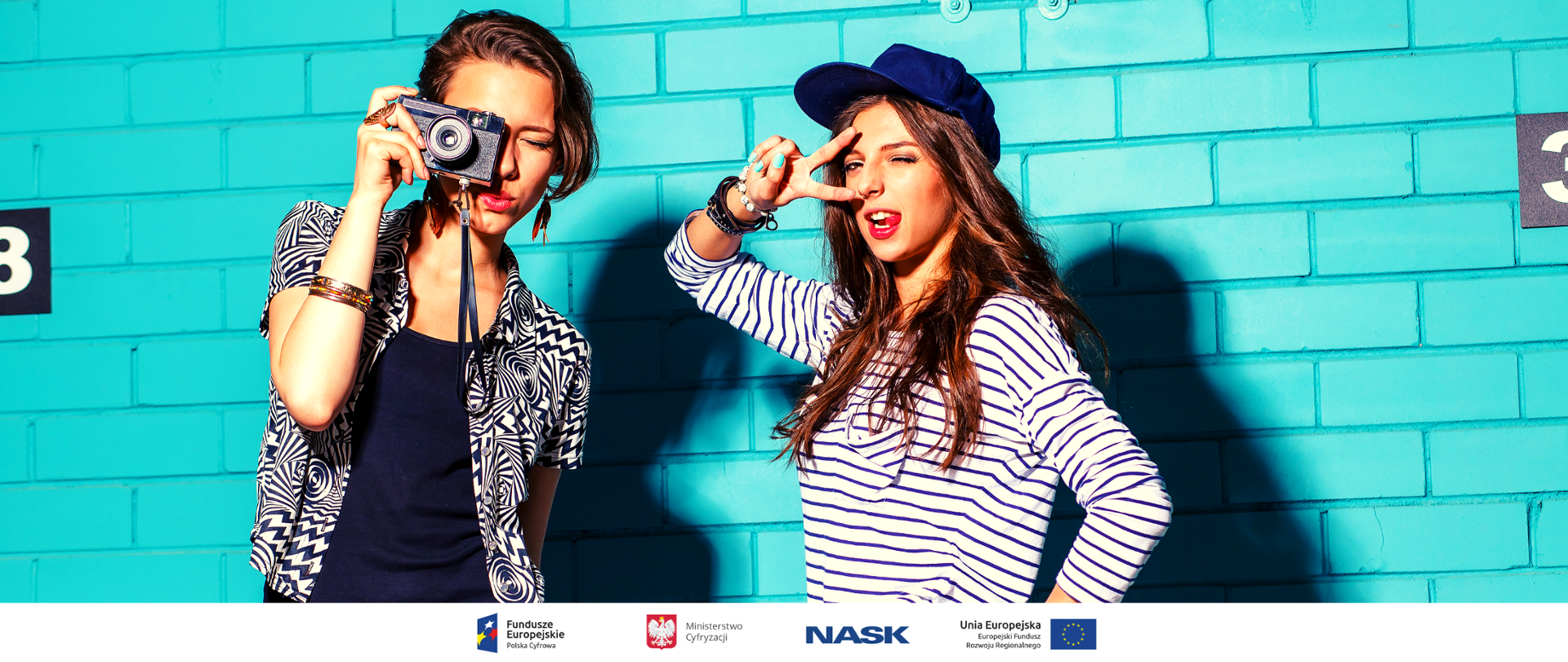 Dwie nastolatki stojące obok niebieskiej ściany, jedna z nich w czapce z daszkiem druga trzyma aparat fotograficzny.