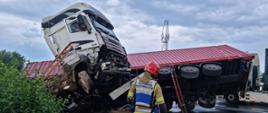 Samochód ciężarowy po wypadku drogowym w wyniku uderzenia leży na prawym boku, część naczepy leży na drodze natomiast przód z ciągnikiem siodłowym są w rowie. Przed samochodem stoi strażak obserwujący miejsce działań. 