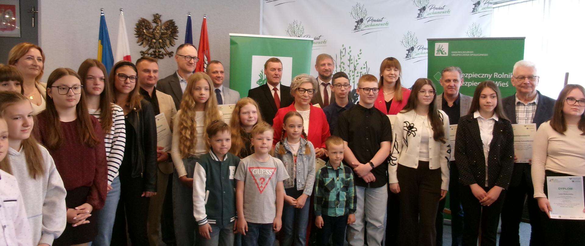 W sali konferencyjnej na pierwszym planie stoi grupa dzieci w różnym wieku, zarówno chłopcy jak i dziewczęta. Za nimi stoją fundatorzy nagród, mężczyźni w garniturach i kobiety, w tym jedna w jaskrawoczerwonym żakiecie. W tle po lewej stronie widać polskie flagi oraz godło Polski. Po prawej stronie znajdują się zielone banery z logo KRUS. 