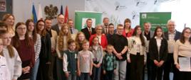 W sali konferencyjnej na pierwszym planie stoi grupa dzieci w różnym wieku, zarówno chłopcy jak i dziewczęta. Za nimi stoją fundatorzy nagród, mężczyźni w garniturach i kobiety, w tym jedna w jaskrawoczerwonym żakiecie. W tle po lewej stronie widać polskie flagi oraz godło Polski. Po prawej stronie znajdują się zielone banery z logo KRUS. 