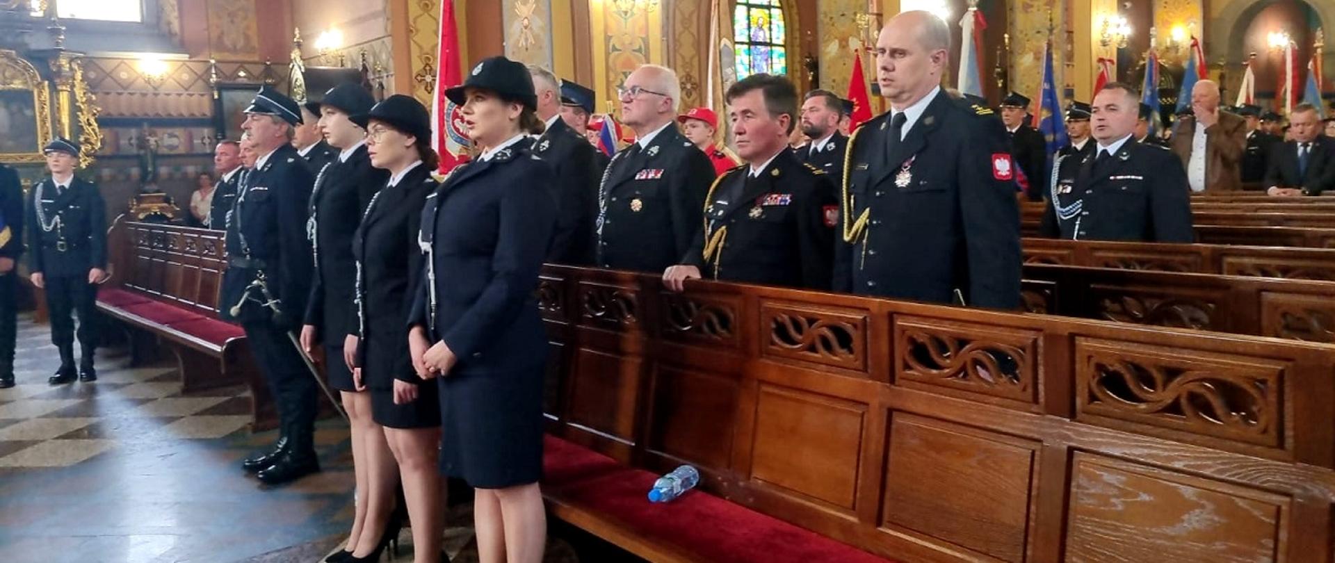 Fotografia wykonana podczas uroczystość 100 lecia istnienia oraz nadanie sztandaru dla jednostki OSP Malinie I. Na fotografii podczas Mszy Świętej stoją w kolejności: zastępca podkarpackiego komendanta wojewódzkiego Państwowej Straży Pożarnej st. bryg. Andrzej Marczenia, komendant powiatowy Państwowej Straży Pożarnej w Mielcu mł. bryg. Łuasz Kapinos, prezes powiatowy związku ochotniczych straży pożarnych w Mielcu druh Franciszek Augustyn. Przed strażakami stoją trzy druhny. Wszyscy ubrani w mundury galowe koloru granatowego. W tle widać liczne sztandary jednostek ochotniczych straży pożarnych z gminy Tuszów Narodowy. Zdjęcie wykonane wewnątrz Kościoła.
