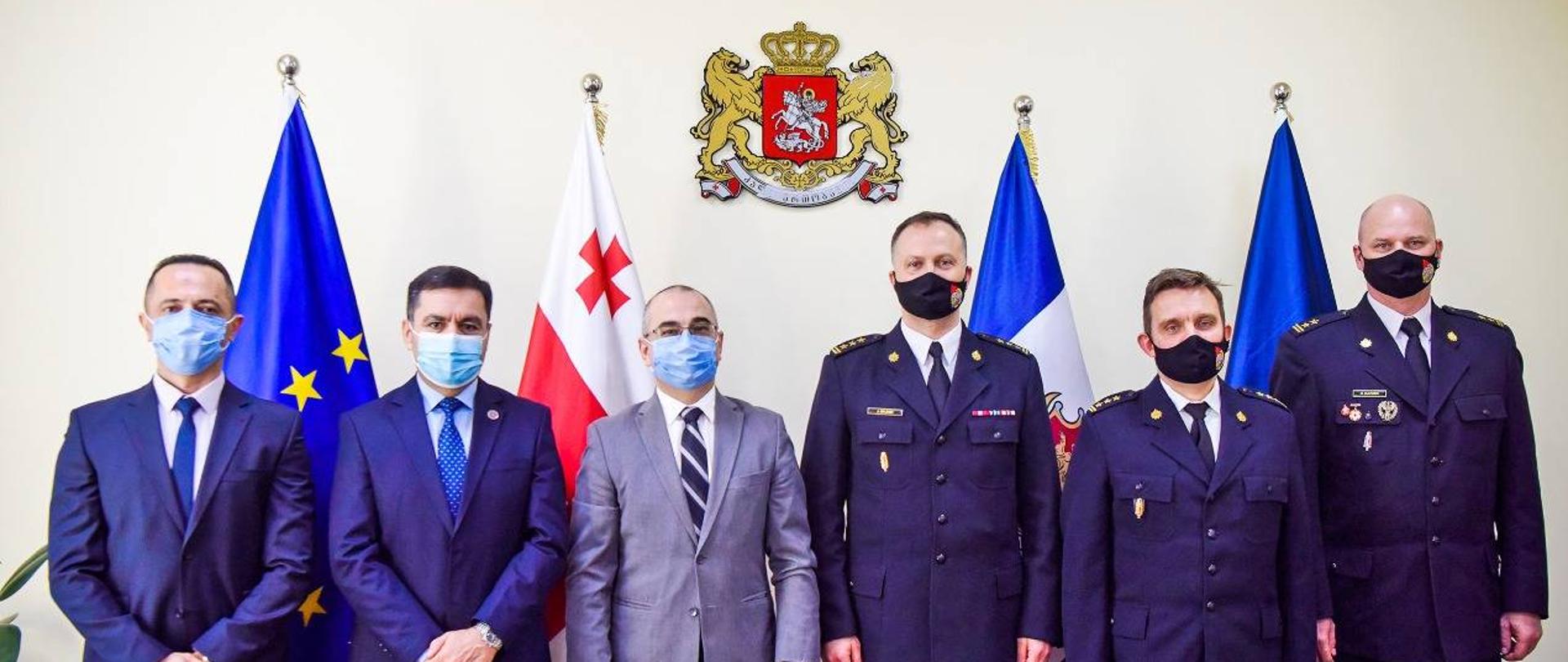 Zdjęcie pamiątkowe uczestników. Trzech mężczyzn w garniturach oraz trzech strażaków Państwowej Straży Pożarnej. Wszyscy w maseczkach ochronnych W tle godło oraz flagi