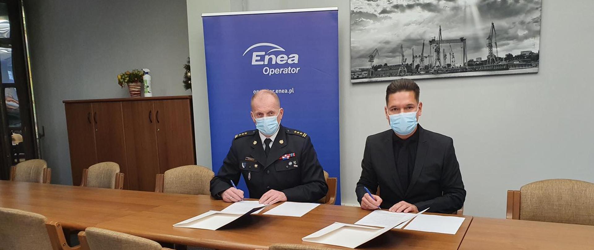 Zachodniopomorski Komendant Wojewódzki Państwowej Straży Pożarnej st. bryg. Jarosław Tomczyk z wiceprezesem ENEA Operator zawierający umowę. W tle baner ENEA operator, panorama stoczni szczecińskiej.