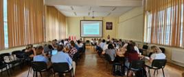 Na zdjęciu na sali konferencyjnej duża grupa osób siedząca przy stołach w oddali zdjęcia tablica z prezentacją