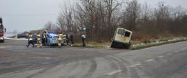 Kolizja dostawczaków. Droga gminna w Dębnie Polskim. Przy wjeździe do jednej z firm, częściowo w rowie znajduje się samochód dostawczy, uczestniczący w kolizji. Z lewej strony z włączonymi sygnałami błyskowymi stoi radiowóz policyjny. Wokół niego są strażacy, policjant oraz uczestnicy zdarzenia. W tle krzaki.