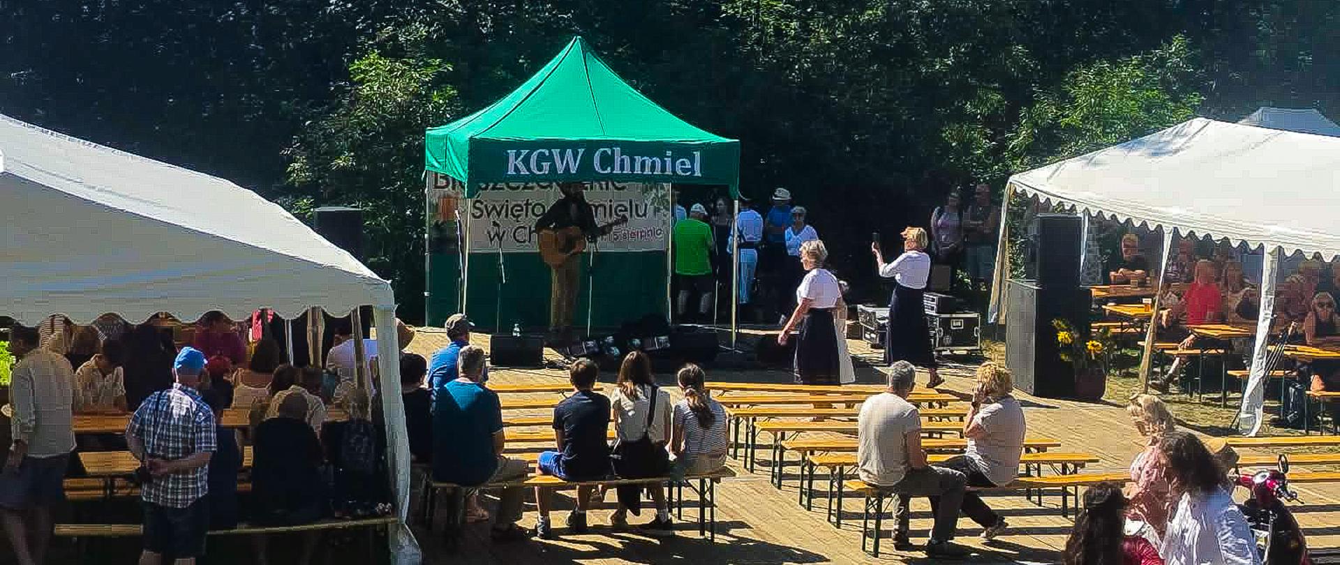  Plenerowe wydarzenie wśród drzew. Na środku znajduje się mała scena z zielonym namiotem oznaczonym „KGW Chmiel” i napisem „Święto Chmielu” Przed sceną ustawiono drewniane ławki, na których siedzą uczestnicy. Po obu stronach znajdują się białe namioty z kolejnymi stoiskami i osobami. W tle widać gęsty las, a nad nim błękitne niebo z chmurami.
