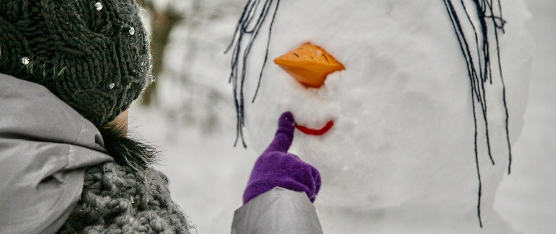 dziewczynka w zimowej kurtce, czapce i rękawiczkach stoi tyłem, ale przodem do twarzy śniegowego bałwana i poprawia mu uśmiech z czerwonej nitki. 