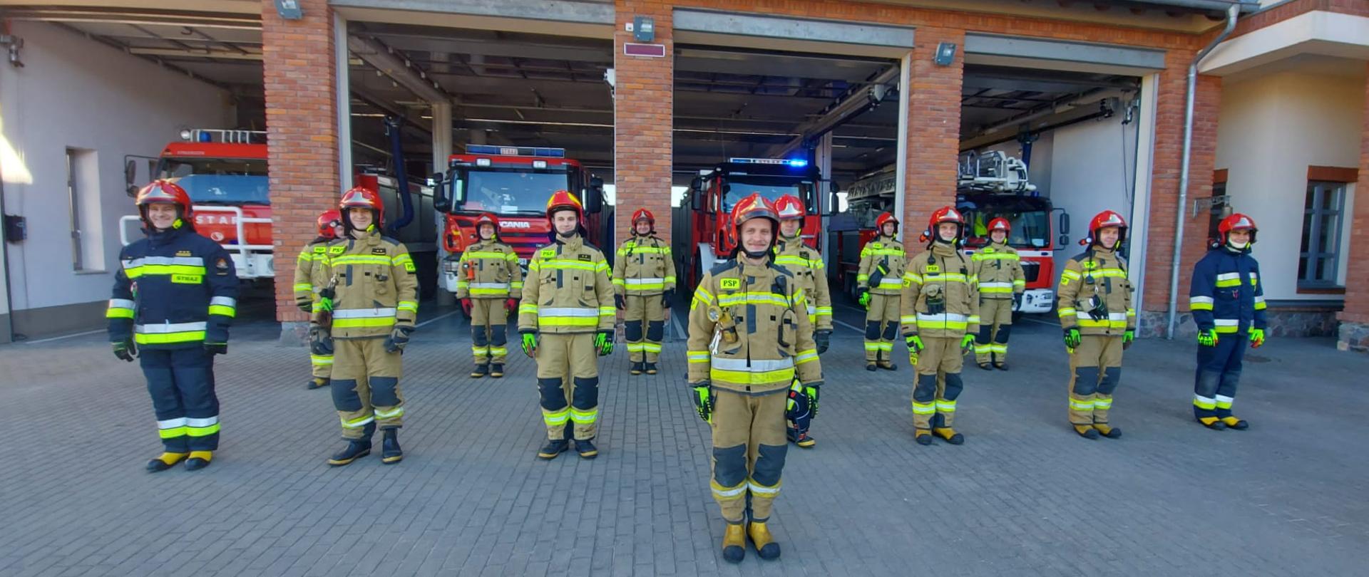 Zdjęcie przedstawia trzynastu strażaków Komendy Powiatowej Państwowej Straży Pożarnej w Gołdapi na tle czterech pojazdów pożarniczych zlokalizowanych w garażu komendy, przed przystąpieniem do wykonania dziesięciu pompek w ramach akcji charytatywnej dla KINGI
