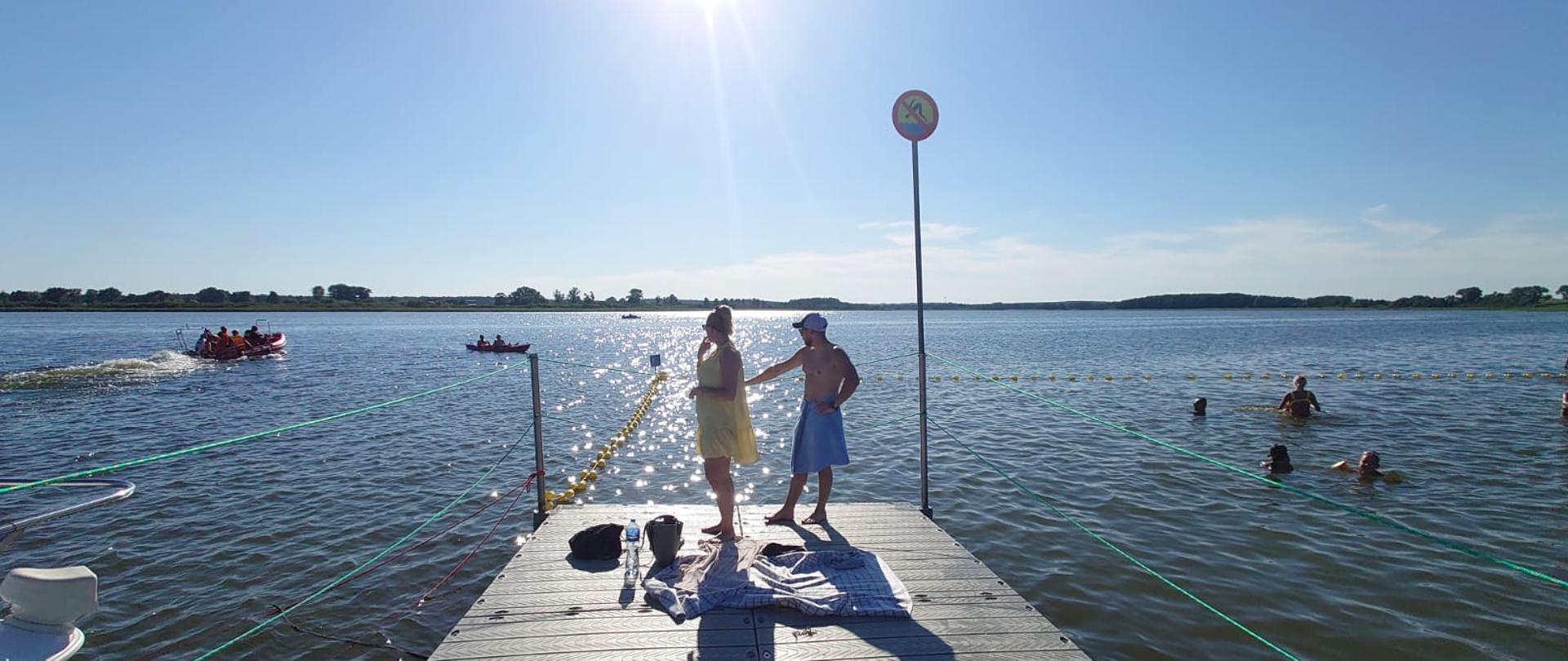 Widok na pomost. Na pomoście stoją 2 osoby. Kobieta ubrana na żółto i mężczyzna owinięty w pasie niebieskim ręcznikiem. Po lewej stronie na wodzie pływają strażacy łodzią ratowniczą. Po prawej stronie w wodzie kąpią się turyści