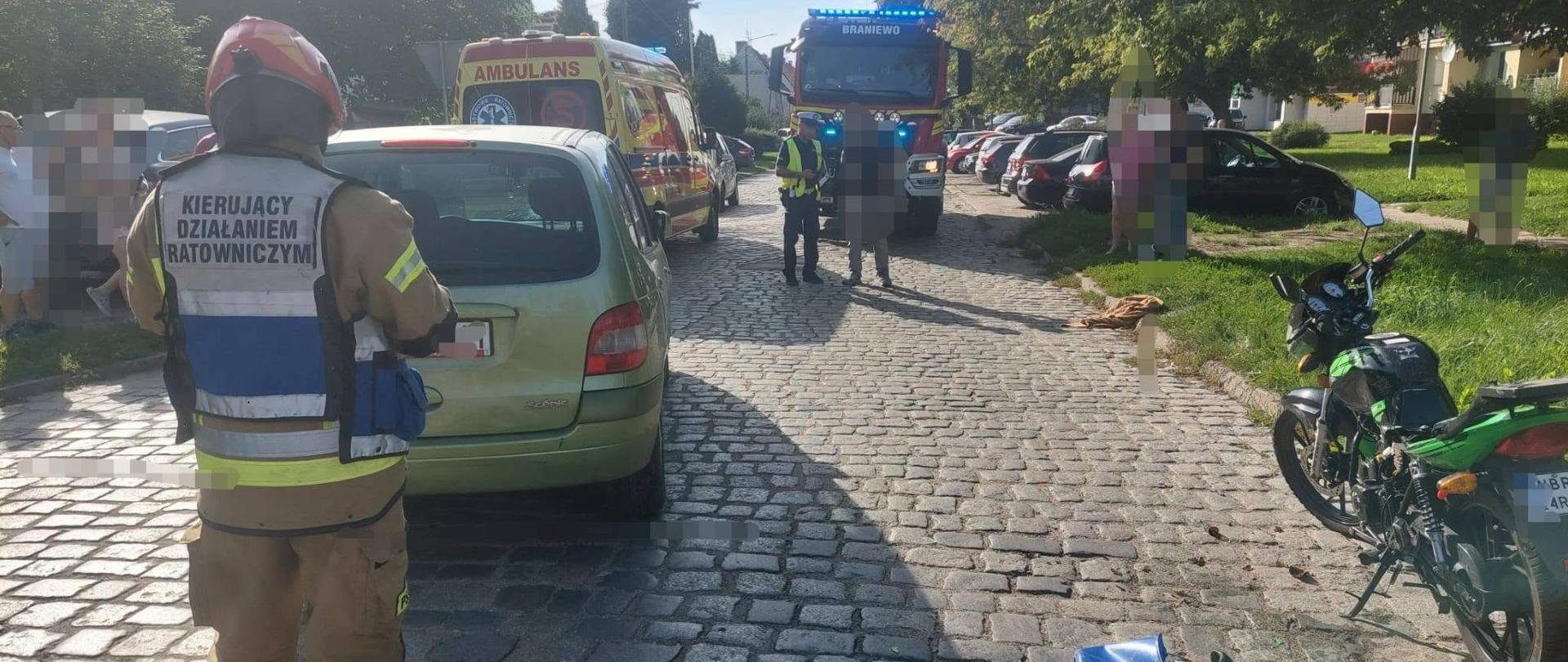 Na zdjęciu widać akcję ratowniczą po zdarzeniu z udziałem motocykla. Na miejscu pracują strażacy i ratownicy medyczni, a teren jest zabezpieczany. Widoczny jest również samochód osobowy oraz uszkodzony motocykl.