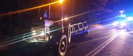 Na zdjęciu w porze nocnej na drodze krajowej numer dwadzieścia siedem doszło do kolizji drogowej ciągnika rolniczego z przyczepom i samochodu osobowego. wyniku najechania na przyczepę samochód osobowy dachował i znalazł się po za drogą. na drodze ustawione są również samochody straży pożarnej.