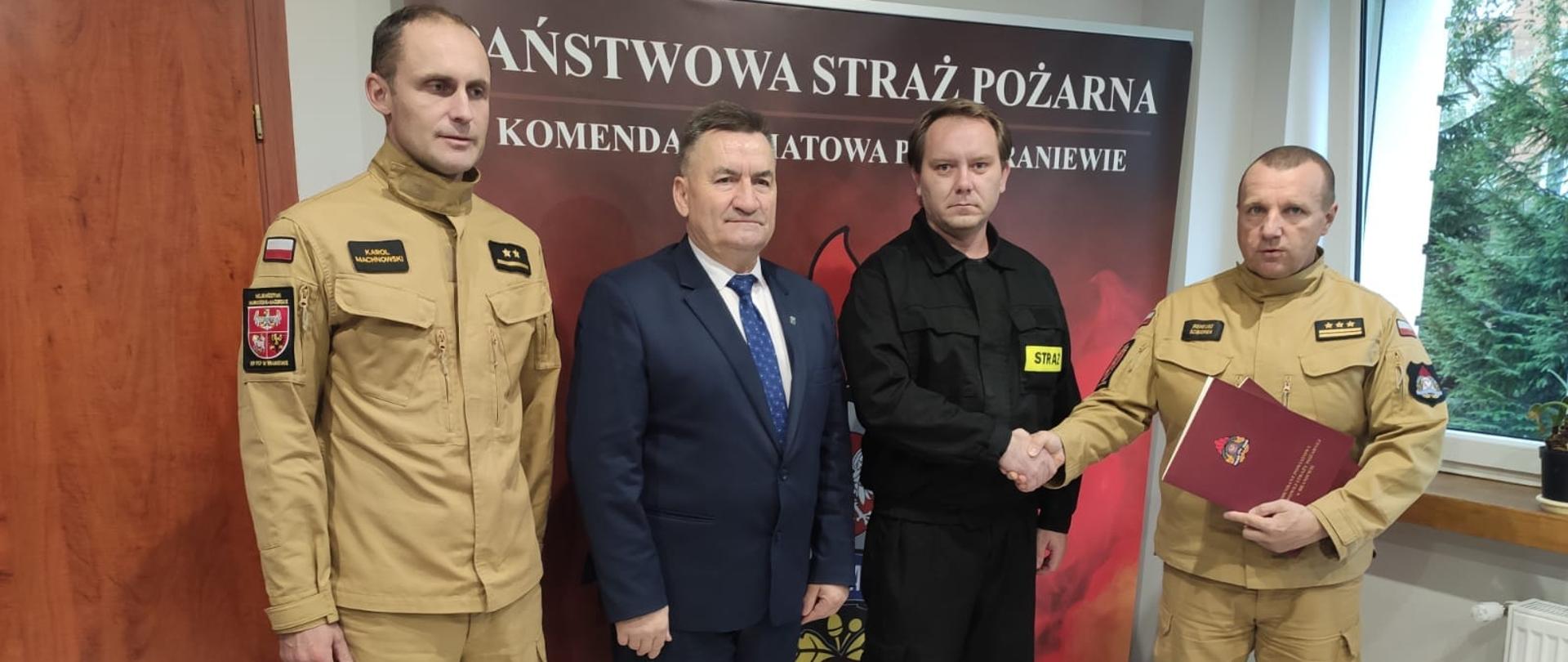 Zdjęcie zrobione w biurze komendanta powiatowego. Od prawej stoją komendant Powiatowy, strażak ochotnik, wice starosta i naczelnik wydziału operacyjnego.