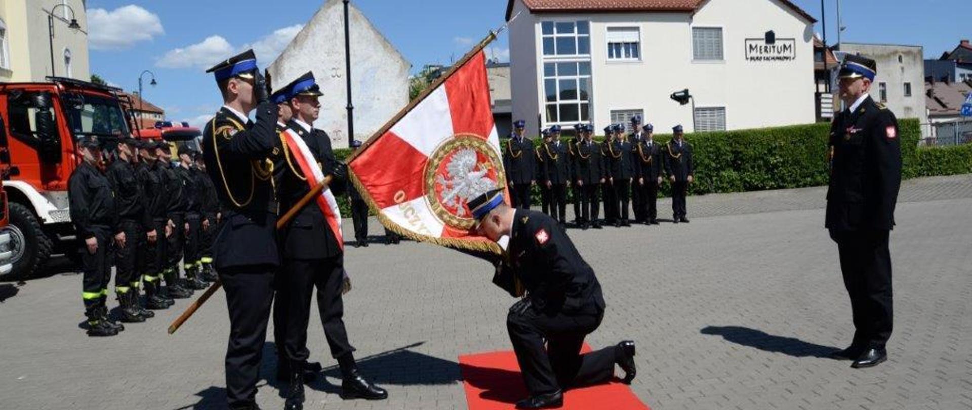 Na zdjęciu widać funkcjonariuszy Komendy Powiatowej PSP w Tucholi w mundurach wyjściowych. Na środku na czerwonym dywanie klęczy Komendant Powiatowy PSP w Tucholi i całuje sztandar. Na sztandarze widać orła białego. Sztandar jest w kolorze biało czerwonym. Przed Komendantem stoi trzech strażaków z pocztu sztandarowego. Strażak po środku trzyma sztandar. Za Komendantem Powiatowym PSP w Tucholi stoi Kujawsko-Pomorski Komendant Wojewódzki PSP. W oddali widać strażaków ustawionych w rzędzie. Za strażakami widać żywopłot i budynki. Po lewej stronie za pocztem sztandarowym stoją strażacy ustawieni w rzędzie. Za strażakami widać wozy strażackie. Wozy są koloru czerwonego. Strażacy stoją na placu.