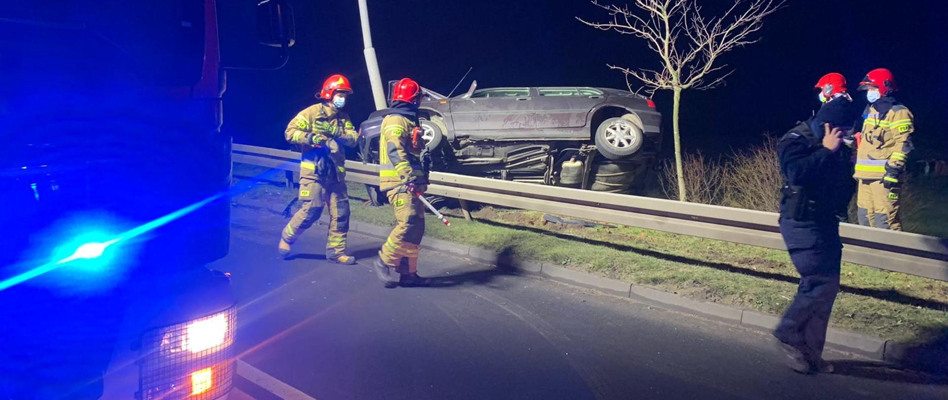 Na zdjęciu widać rozbite auto oraz strażaków i policjantów wykonujących czynności ratowniczo-dochodzeniowe 