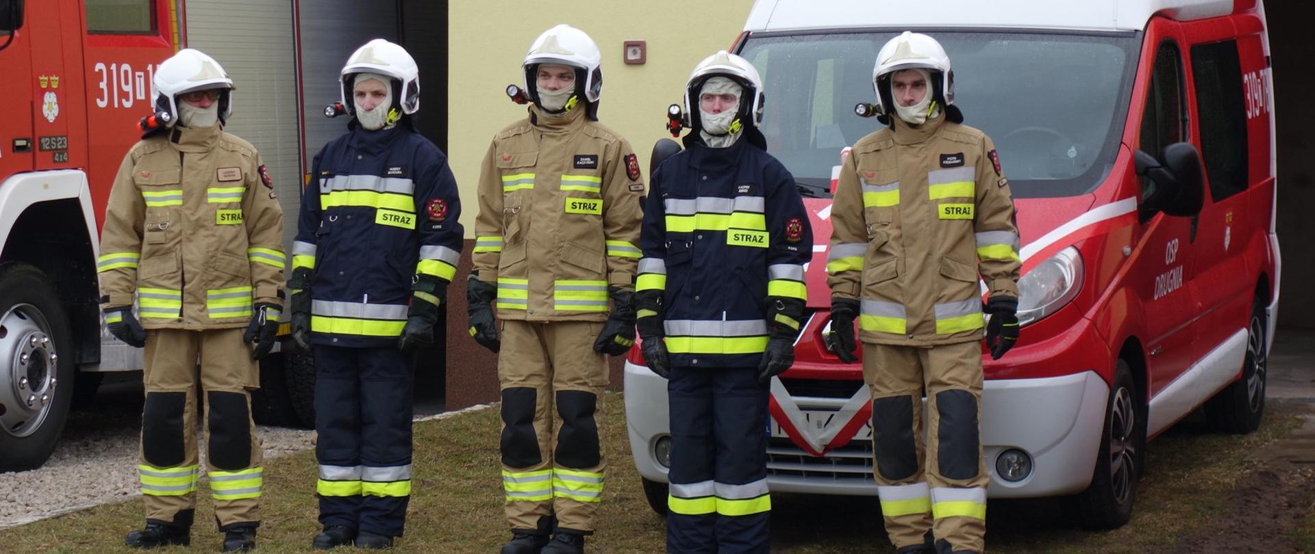 Na zdjęciu widzimy grupę pięciu strażaków stojących na tle samochodu pożarniczego