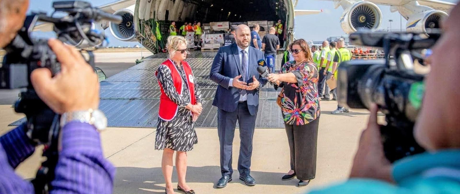 Na płycie lotniska trwa briefing prasowy przy otwartej rampie dużego samolotu transportowego, z którego rozładowywane są palety z pomocą humanitarną.