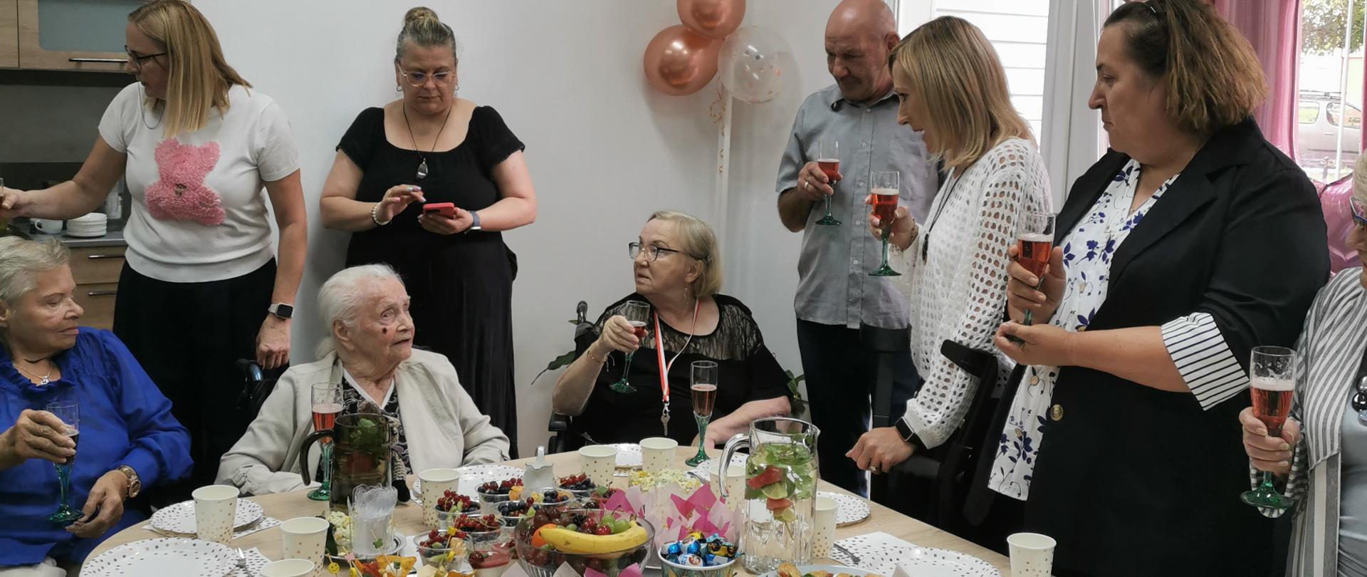 Przy stole z poczęstunkiem, obok ubranej w jasnoszarą marynarkę Jubilatki siedzą na wózkach inwalidzkich starsze pani – koleżanki z DPS Magnolia w Głogowie. Za Jubilatką stoją jej wnuczki, a przy oknie, ozdobionym różowymi balonami z napisem Happy Birthday, od lewej strony stoi mężczyzna w szarej koszuli - Sołtys wsi Grochowice Rober Duda, następnie kobieta w białej bluzce - Wójt Gminy Kotla Kamila Suchocka-Szperlik.
