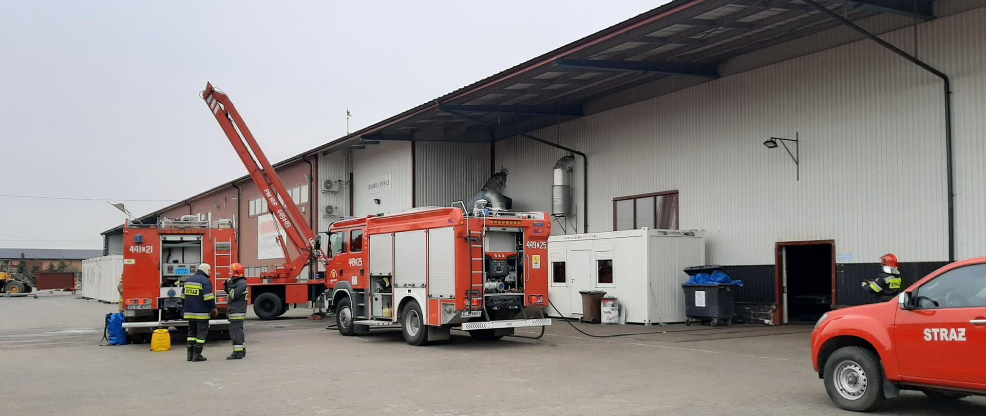 Na zdjęciu widać samochody strażackie oraz strażaków biorących udział w akcji gaśniczej. W tle widać halę produkcyjną, w której doszło do pożaru.