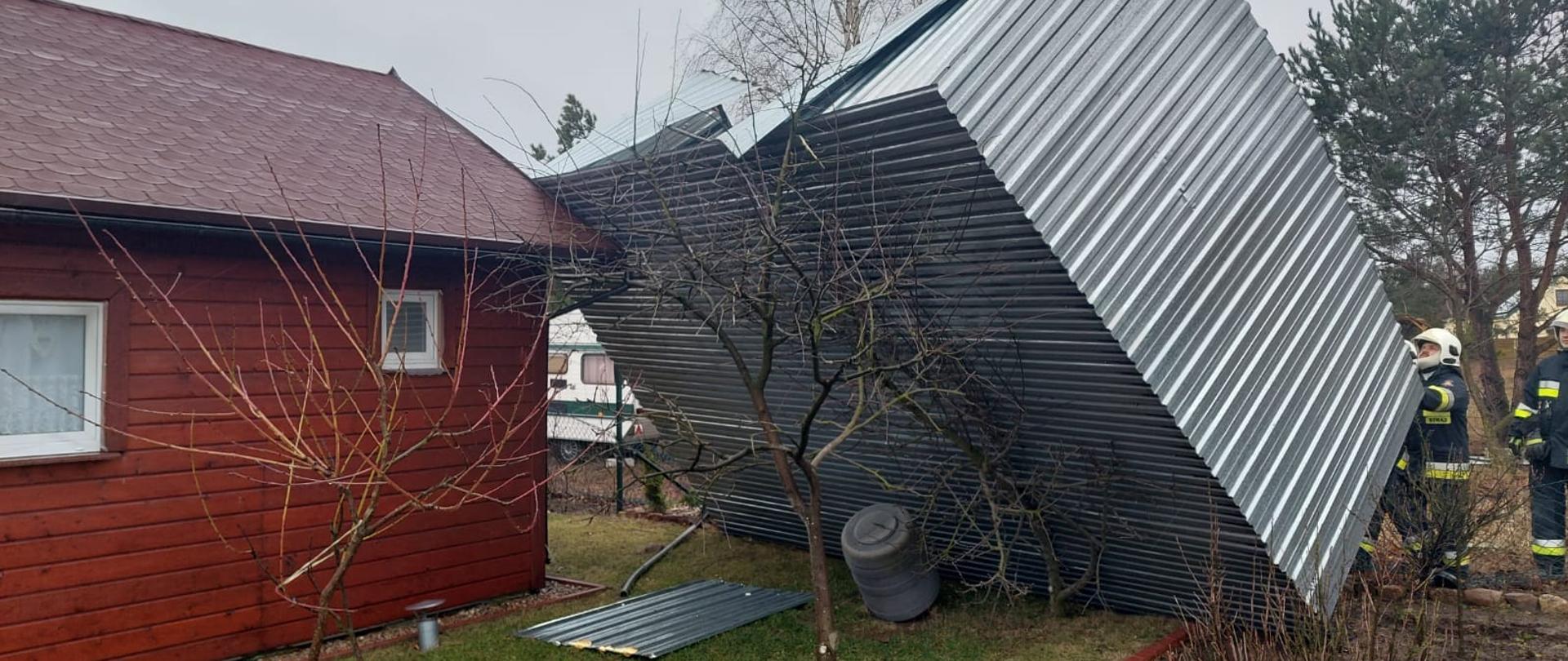 Pochmurno. Duży garaż z blachy falistej w kolorze srebrnym opart podłogą o krawędź brązowego domku letniskowego. Obok stojący strażacy w ciemnych mundurach.
