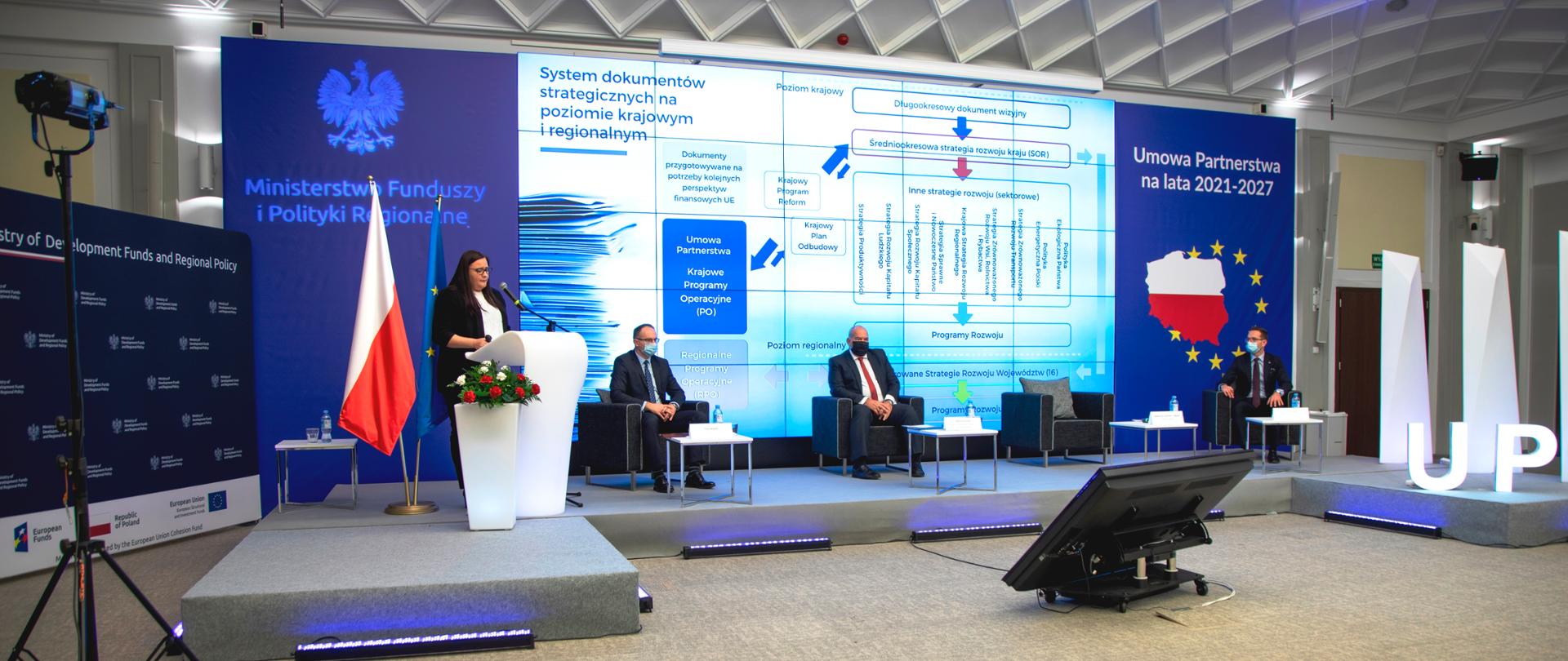 Na sali konferencyjnej na podeście w mównicy stoi wiceminister Małgorzata Jarosińska-Jedynak. Trzech mężczyzn siedzi w fotelach. Od lewej prowadzący spotkanie, minister Tadeusz Kościński i wiceminister Waldemar Buda. Z tyłu na ekranie wyświetlona jest prezentacja.