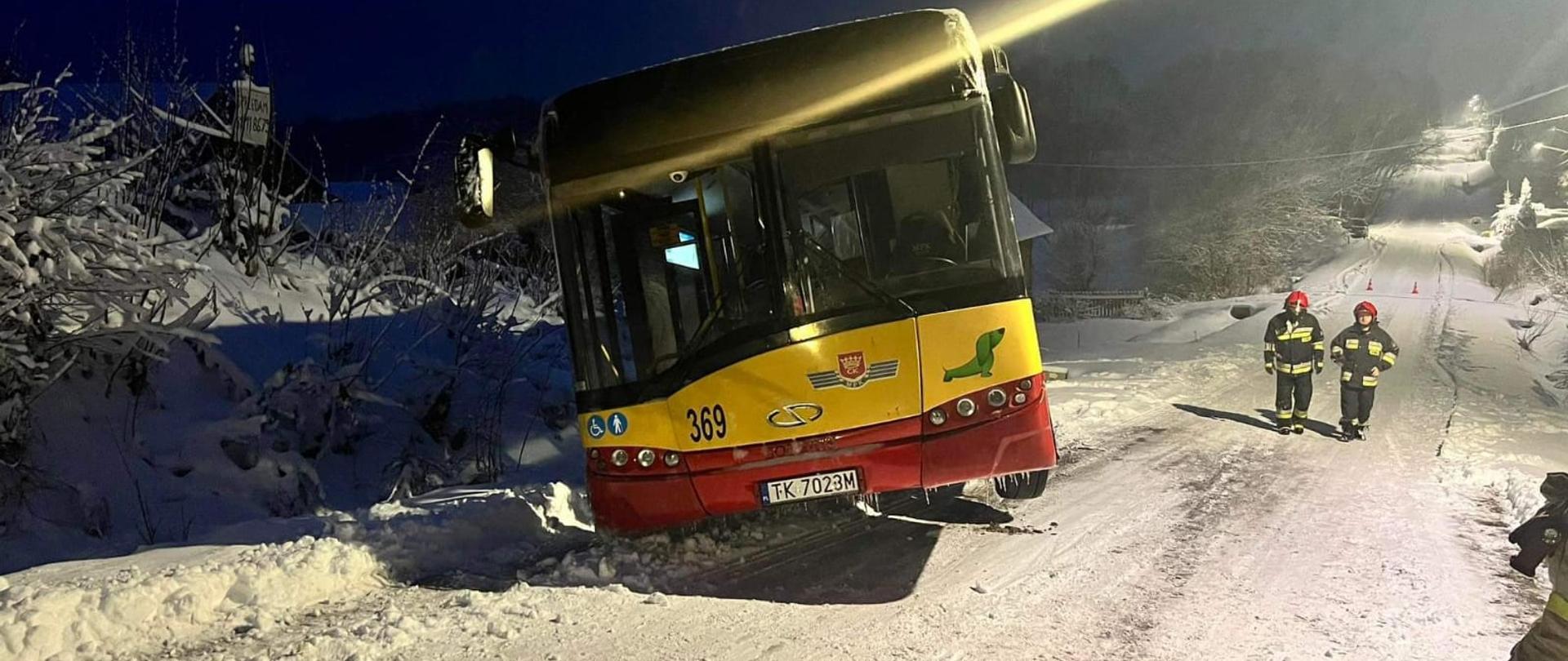 Autobus MZK Kielce wpadł do przydrożnego rowu z powodu śliskiej drogi pokrytej śniegiem i lodem. Tył autobusu znajduje się w rowie, a przód jest lekko uniesiony ponad drogę. Pod górkę w kierunku autobusu idą dwaj strażacy.