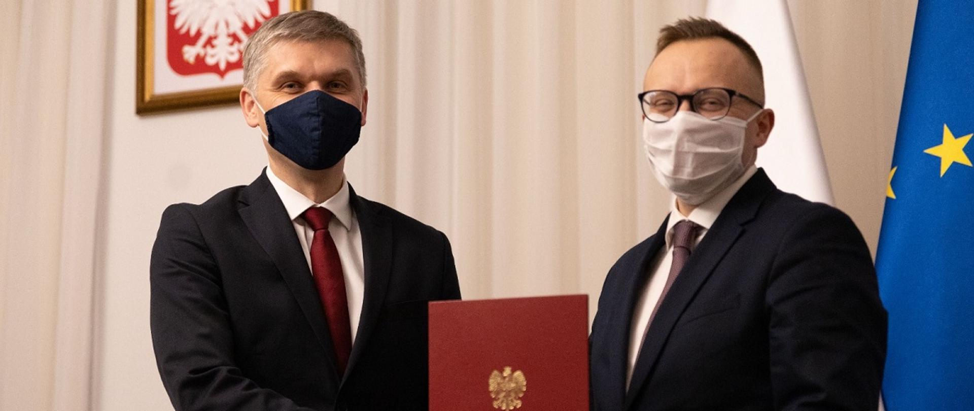 Minister rozwoju i technologii Piotr Nowak ściska rękę Artura Sobonia, który w drugiej dłoni trzyma akt odwołania z funkcji sekretarza stanu w Ministerstwie Rozwoju i Technologii, a także Pełnomocnika Prezesa Rady Ministrów do Spraw Rozwoju Lokalnego. Z tyłu za ich plecami godło Polski oraz flagi Polski i UE.
