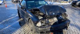 Zdjęcie przedstawia rozbity przód pojazdu osobowego marki BMW stojącego na środku skrzyżowania.
