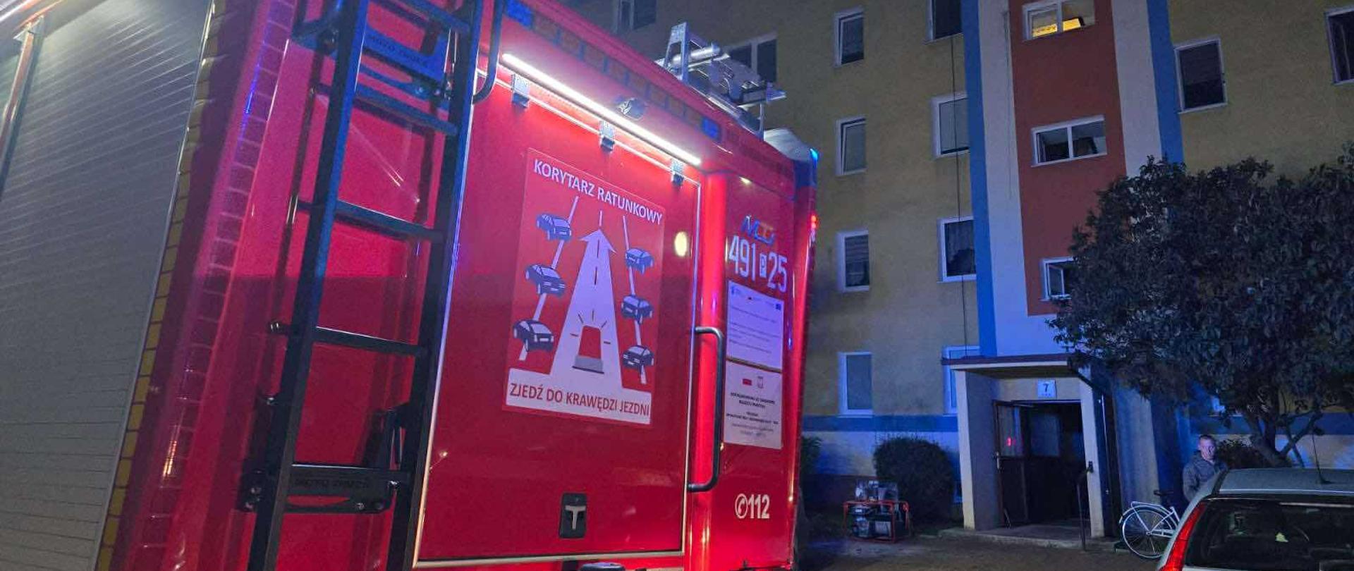 Zdjęcie przedstawia tył zabudowy pojazdu pożarniczego. Na tylnej rolecie widnieje naklejka przedstawiająca zasady zachowania się na drodze tzw. KORYTARZ ŻYCIA na drugim planie elewacja budynku wielorodzinnego.