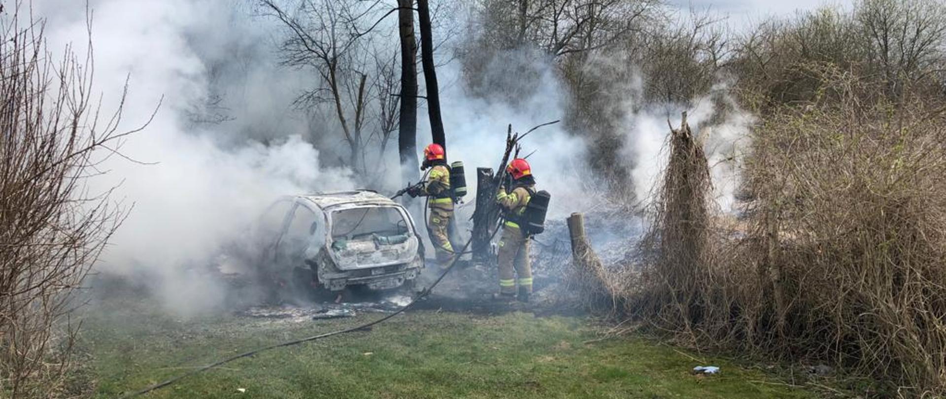 Strażacy gaszą samochód osobowy. Są zabezpieczeni w aparaty ochrony dróg oddechowych. Z pojazdu wydobywa się dym