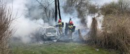 Strażacy gaszą samochód osobowy. Są zabezpieczeni w aparaty ochrony dróg oddechowych. Z pojazdu wydobywa się dym.