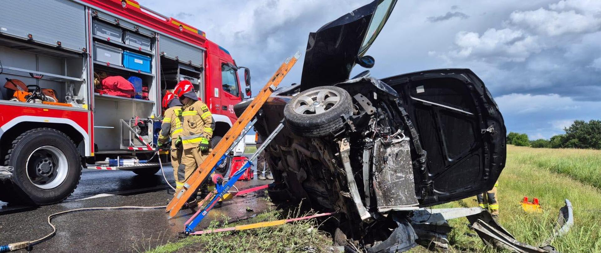 Zdjęcie przedstawia uszkodzony samochód po dachowaniu na Autostradzie A4 oraz pojazdy jednostek ochrony przeciwpożarowej.