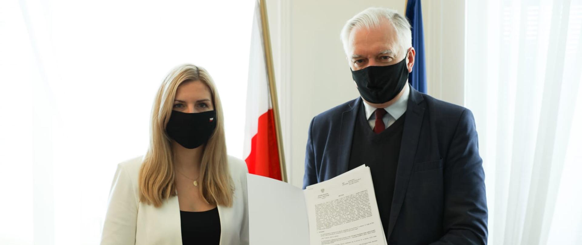 
Wicepremier, minister rozwoju, pracy i technologii Jarosław Gowin w maseczce na twarzy, trzyma w dłoniach dokument, po jego lewej stronie stoi wiceminister rozwoju, pracy i technologii Anna Kornecka. Z tyłu flagi Polski i UE.