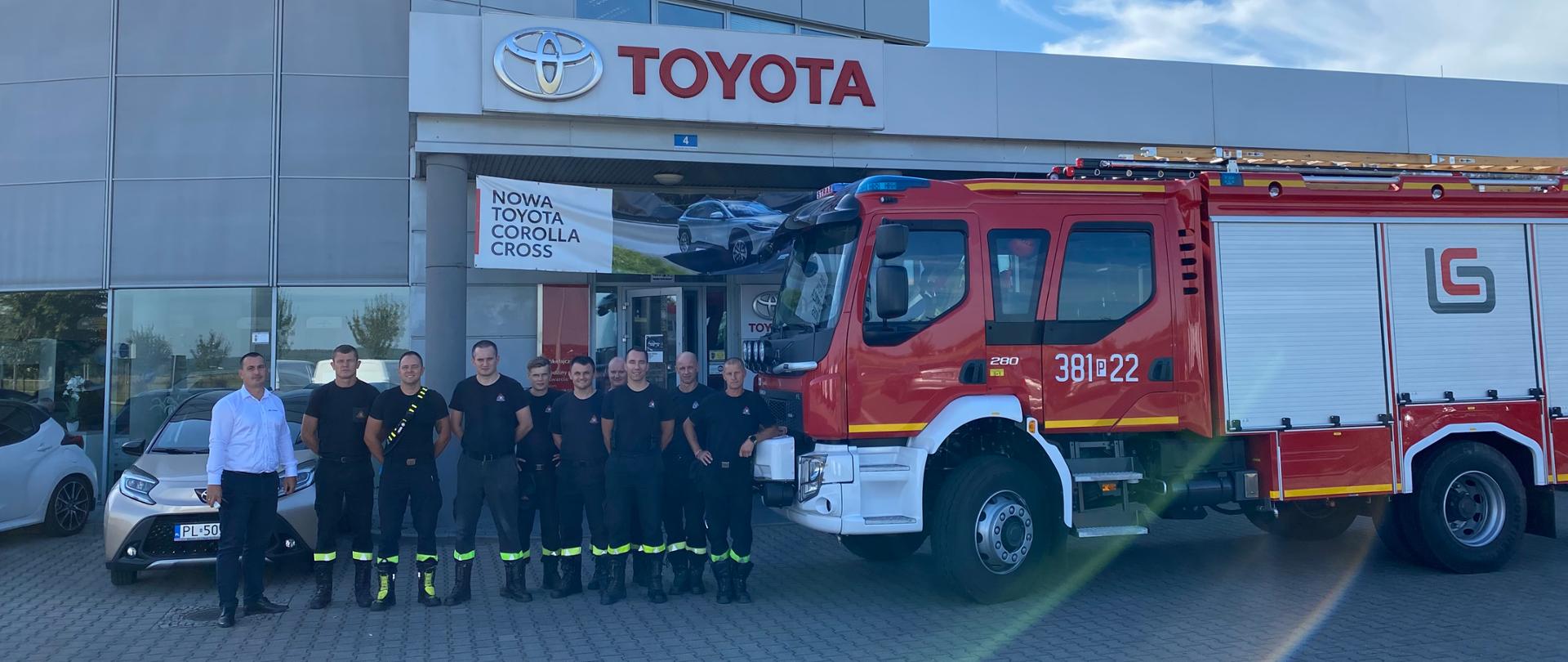 Strażacy i osoba cywilna stoją w dwuszeregu przed salonem samochodowym. Obok stoją samochody i wóz strażacki.